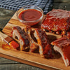 A wooden cutting board displays barbecue ribs covered in sauce, accompanied by a bowl of sauce and a brush.