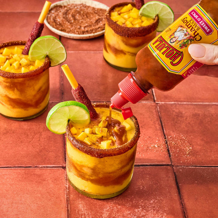A hand pours Cholula Chamoy Sauce into a mango drink with a chili-rimmed glass and a lime wedge.