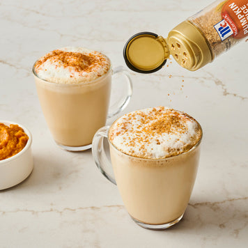 Two glasses of pumpkin spice latte with a bottle of pumpkin spice mix being poured over them on a marble surface.