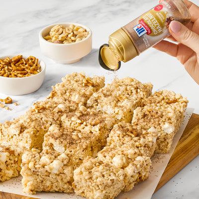 Hand sprinkling McCormick Chocolate Hazelnut Finishing Sugar over popcorn and pretzel snack bars with bowls of peanuts and pretzels in the background