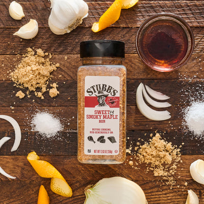 A jar of Stubb's Sweet and Smoky Maple Rub is surrounded by ingredients like garlic, onion, brown sugar, salt, and orange peel on a wooden surface.