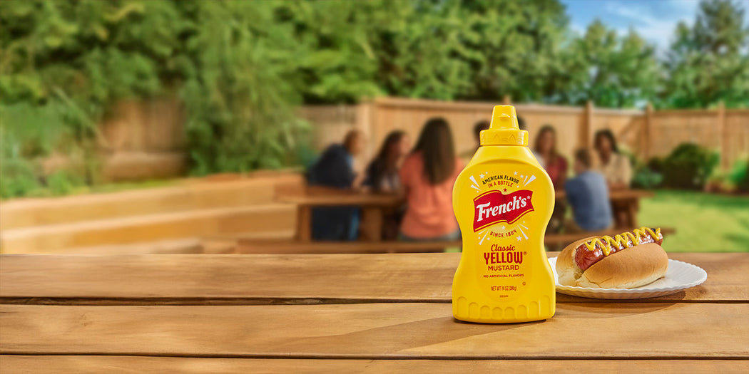 french's classic yellow mustard next to a hot dog with mustard on a picnic table with a family in the background