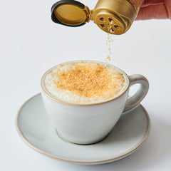 Hand sprinkling finishing sugar over a frothy cappuccino in a white cup and saucer