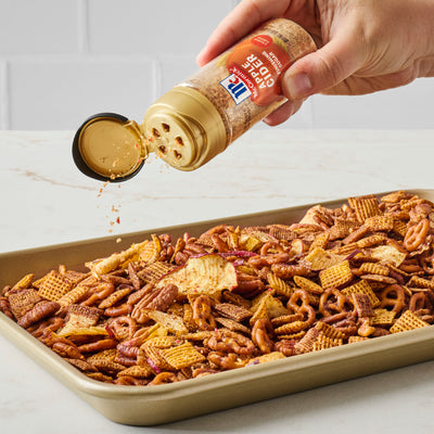 Hand sprinkling seasoning over a tray of snack mix on a white surface