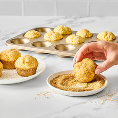 Hand dipping freshly baked muffin into English toffee finishing sugar with more sugar-coated muffins and a muffin tin in the background