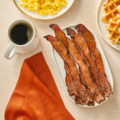 Plate of bacon with a cup of coffee and waffles on a beige tablecloth.