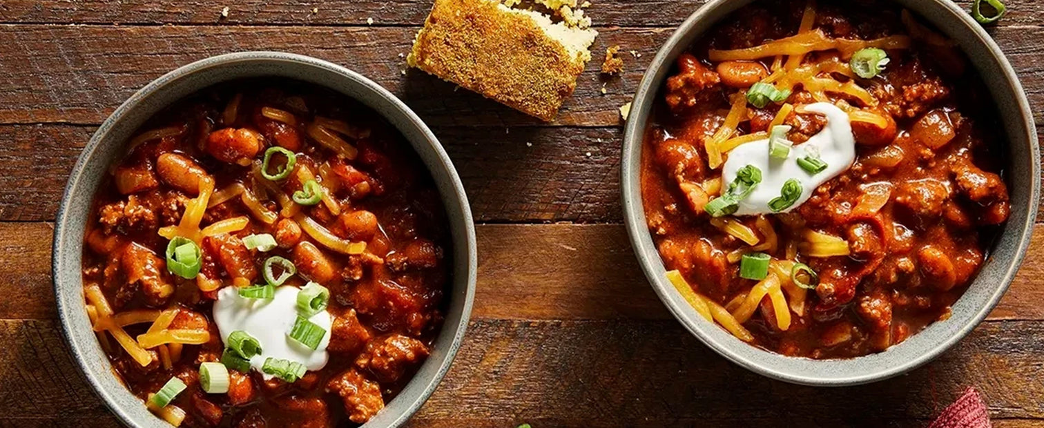 bowl of chili with corn bread, green onions, sour cream, and cheese on a wooden table