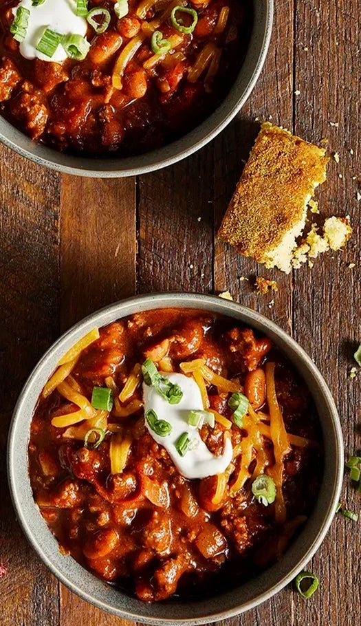 bowl of chili with corn bread, green onions, sour cream, and cheese on a wooden table