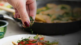 Person garnishing a dish with herbs over a blurred background of a frying pan and ingredients.
