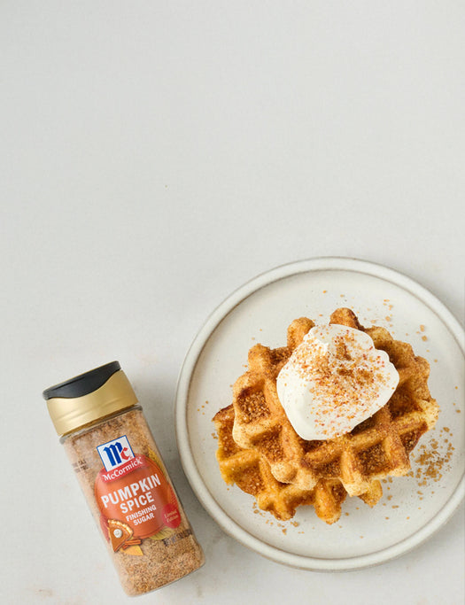 Pumpkin spice seasoning bottle next to a plate of waffles with whipped cream on a light background