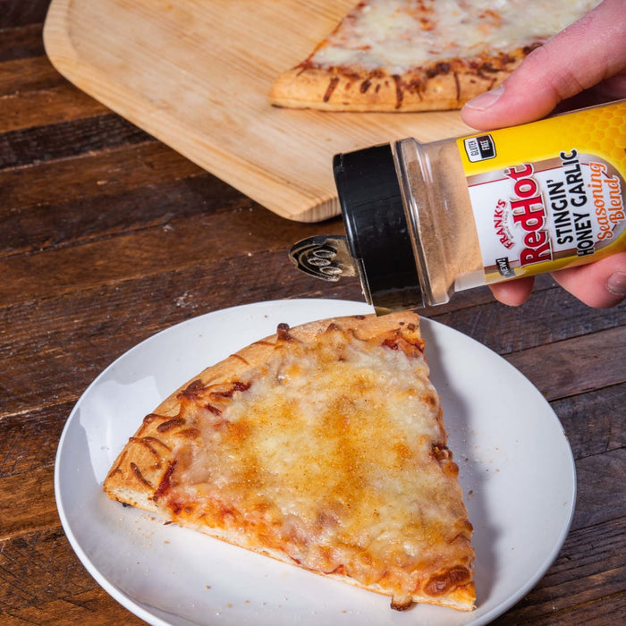 A hand sprinkles Franks RedHot Stingin Honey Garlic Seasoning Blend on a slice of pizza.
