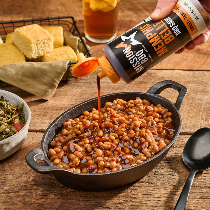 Mission BBQ Memphis Belle BBQ Sauce is poured over a dish of baked beans, served with cornbread and greens.