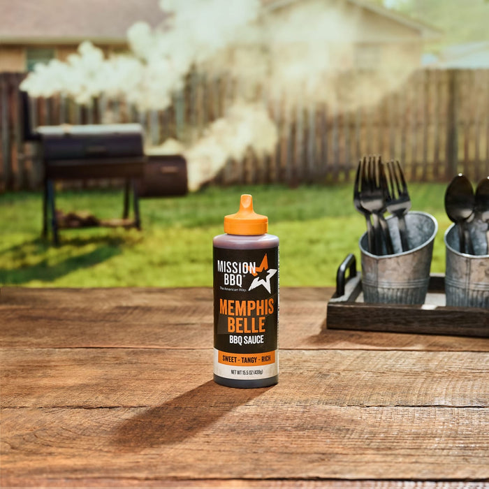 A bottle of Mission BBQ Memphis Belle BBQ Sauce sits on a wooden table with a smoker in the background.