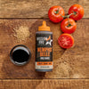 A bottle of Mission BBQ Memphis Belle BBQ Sauce is displayed with tomatoes, mustard seeds, and a bowl of dark liquid on a wooden surface.