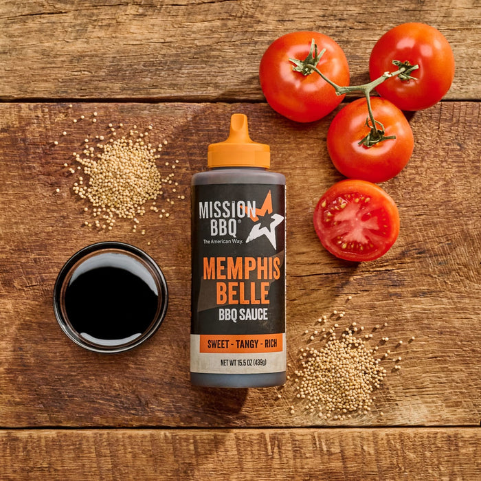 A bottle of Mission BBQ Memphis Belle BBQ Sauce is displayed with tomatoes, mustard seeds, and a bowl of dark liquid on a wooden surface.
