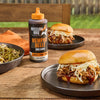 A bottle of Mission BBQ Memphis Belle BBQ Sauce sits on a wooden table next to two pulled pork sandwiches topped with coleslaw and a bowl of green beans.