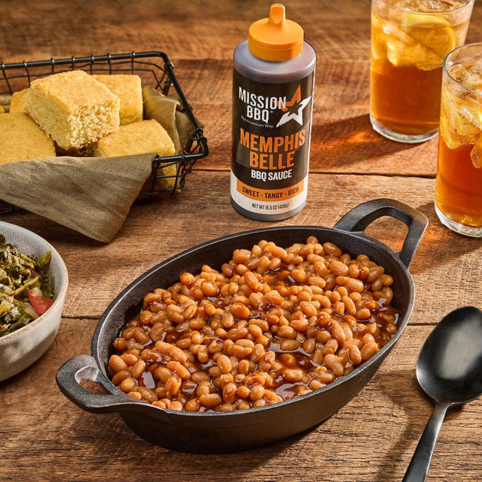 A bottle of Mission BBQ Memphis Belle BBQ Sauce sits on a wooden table next to a cast iron dish of baked beans, cornbread, collard greens, and iced tea.