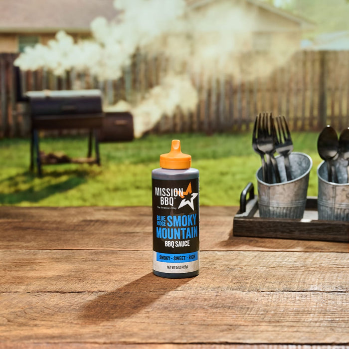 A bottle of Mission BBQ Blue Ridge Smoky Mountain BBQ Sauce sits on a wooden table with a smoker in the background.