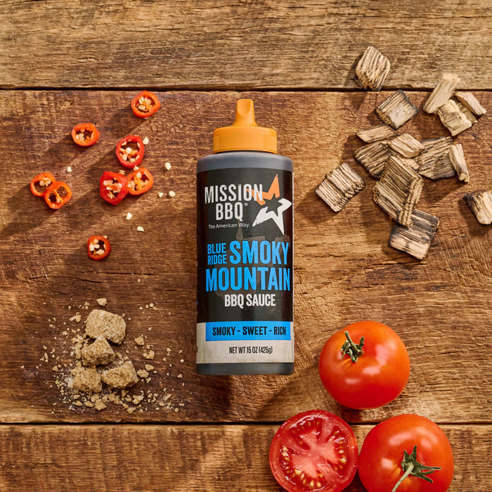 A bottle of Mission BBQ Blue Ridge Smoky Mountain BBQ Sauce is displayed on a wooden surface with ingredients including tomatoes, peppers, brown sugar, and wood chips.