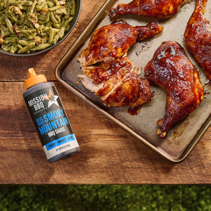 A bottle of Mission BBQ Blue Ridge Smoky Mountain BBQ Sauce sits next to a tray of glazed chicken and a bowl of green beans.