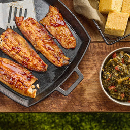 Four glazed fish fillets, coated in a rich sauce like Mission Bbq Blue Ridge Smoky Mountain 15 Oz, are presented in a black cast iron pan alongside a bowl of collard greens and cornbread on a rustic wooden table.