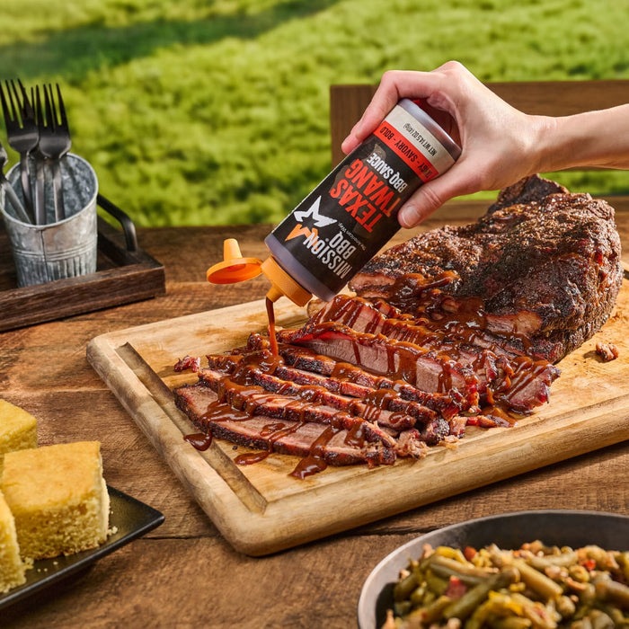 A hand pours Mission BBQ Texas Twang BBQ Sauce onto sliced brisket on a wooden cutting board, accompanied by cornbread and green beans.