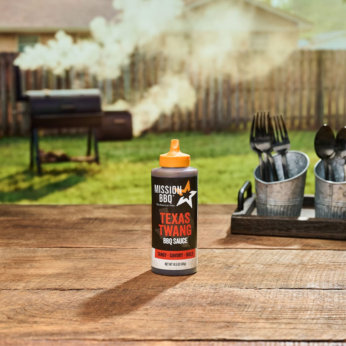 Mission BBQ Texas Twang BBQ Sauce sits on a wooden table with a grill and utensils in the background.