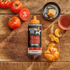 A bottle of Mission BBQ Texas Twang BBQ Sauce is displayed on a wooden surface with tomatoes, honeycomb, and mandarin oranges.