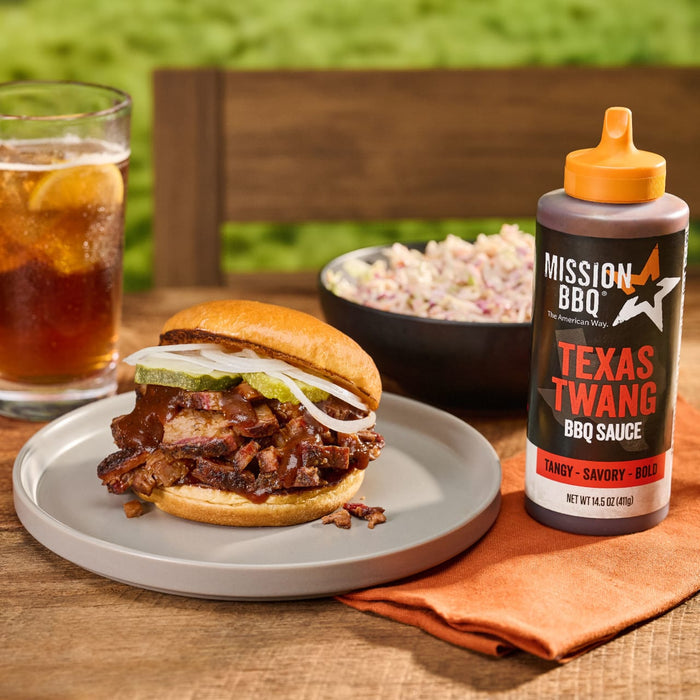 A Mission BBQ Texas Twang BBQ Sauce bottle sits next to a brisket sandwich with coleslaw and a glass of iced tea.