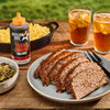 A bottle of Mission BBQ Texas Twang BBQ Sauce sits on a wooden table next to a plate of sliced meatloaf, mac and cheese, collard greens, and two glasses of iced tea.