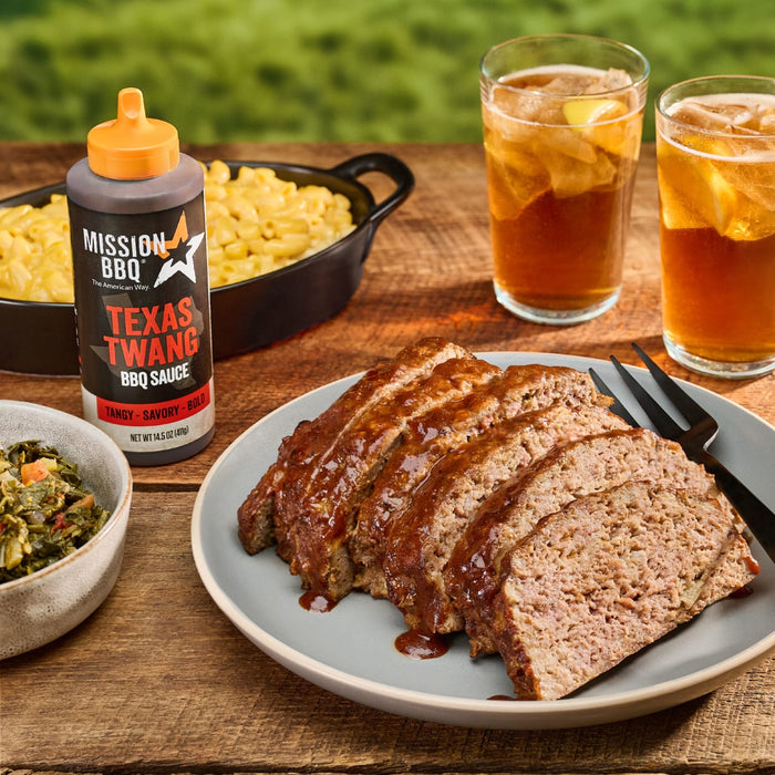 A bottle of Mission BBQ Texas Twang BBQ Sauce sits on a wooden table next to a plate of sliced meatloaf, mac and cheese, collard greens, and two glasses of iced tea.