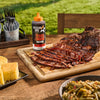 A bottle of Mission BBQ Texas Twang BBQ Sauce sits on a wooden table next to sliced brisket, cornbread, and green beans.