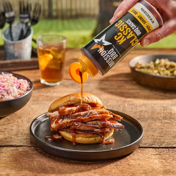 A hand pours Mission BBQ KC Classic BBQ Sauce onto a sandwich filled with sliced meat.