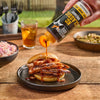 A hand pours Mission BBQ KC Classic BBQ Sauce onto a sandwich filled with sliced meat.