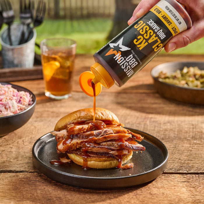 To showcase its rich consistency, a bottle of Mission Bbq Kc Classic Bold Barbecue Sauce Plastic Squeeze Bottle 16 Oz is squeezed over a piled-high meat sandwich on a rustic wooden table.