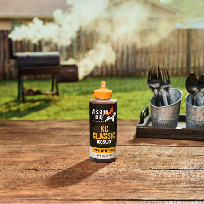 A bottle of Mission BBQ KC Classic BBQ Sauce sits on a wooden table with a smoker in the background.