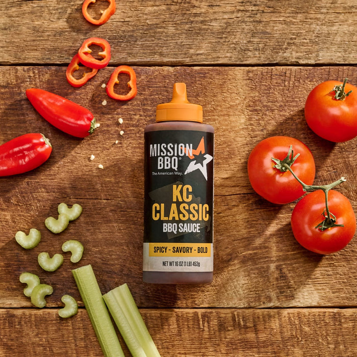A bottle of Mission BBQ KC Classic BBQ Sauce is displayed on a wooden surface with fresh tomatoes, red peppers, and celery.