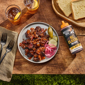 A bottle of Mission BBQ KC Classic BBQ Sauce sits next to a plate of brisket burnt ends with pickled onions and jalapeño, accompanied by drinks and bread.