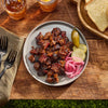 A plate of brisket burnt ends, glazed with Mission Bbq Kc Classic Bold Barbecue Sauce Plastic Squeeze Bottle 16 Oz, is served on a wooden table with pickles, pickled onions, and bread.