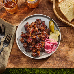 A plate of brisket burnt ends, glazed with Mission Bbq Kc Classic Bold Barbecue Sauce Plastic Squeeze Bottle 16 Oz, is served on a wooden table with pickles, pickled onions, and bread.