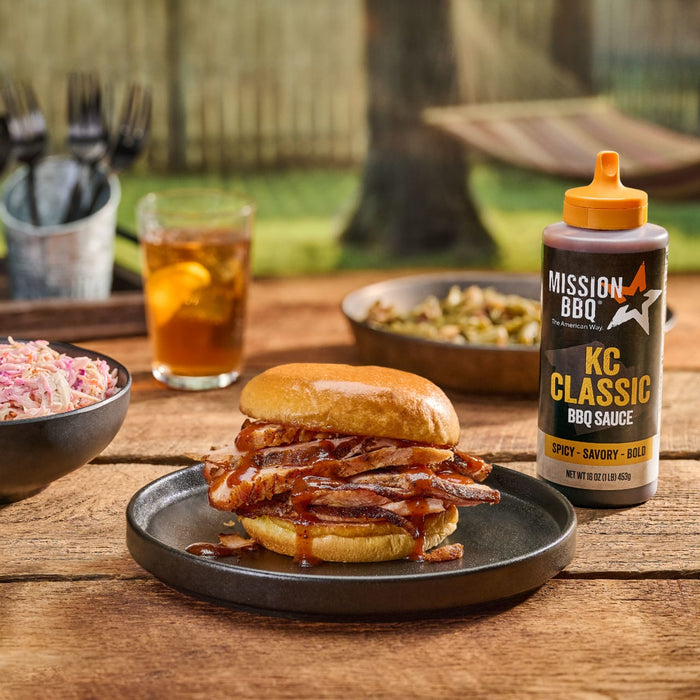 A bottle of Mission BBQ KC Classic BBQ Sauce sits next to a barbecue sandwich on a wooden table with coleslaw and other side dishes, suggesting a backyard barbecue setting.