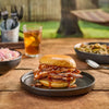 A delicious sliced meat sandwich, generously drizzled with Mission Bbq Kc Classic Bold Barbecue Sauce Plastic Squeeze Bottle 16 Oz, is served on a rustic wooden table with coleslaw and iced tea for a perfect backyard meal.
