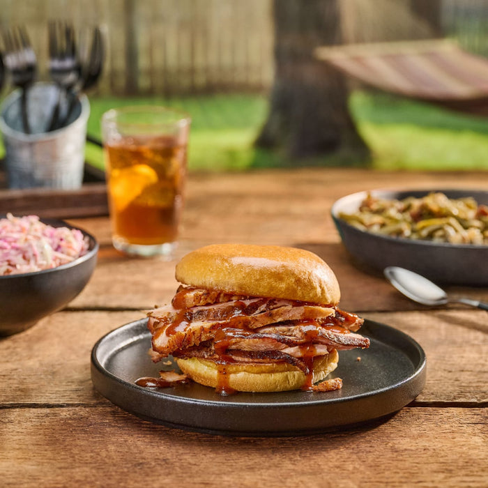 A delicious sliced meat sandwich, generously drizzled with Mission Bbq Kc Classic Bold Barbecue Sauce Plastic Squeeze Bottle 16 Oz, is served on a rustic wooden table with coleslaw and iced tea for a perfect backyard meal.