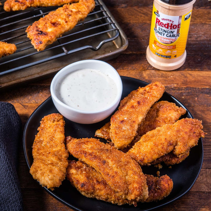 A plate of crispy chicken tenders with a side of ranch dressing and a bottle of Franks RedHot Stingin Honey Garlic Seasoning Blend.