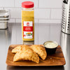 A wooden plate with two golden empanadas and a side of dipping sauce, next to a bottle of Goya Sazon Tropical seasoning.