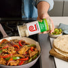 A hand sprinkles Lawry's Garlic Salt over a skillet of sizzling fajita ingredients.