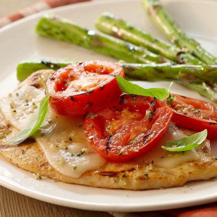A plate features grilled chicken topped with melted cheese and grilled tomato slices, garnished with basil, alongside grilled asparagus.