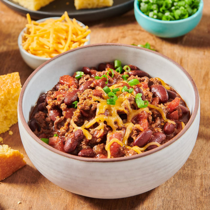 A bowl of chili with kidney beans, ground beef, tomatoes, and shredded cheddar cheese, garnished with green onions, served with cornbread and additional toppings.