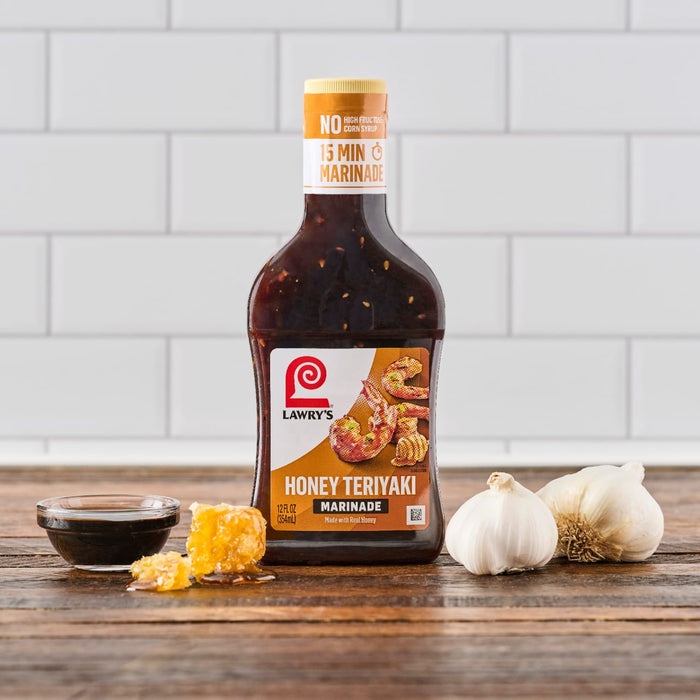 A bottle of Lawrys Honey Teriyaki Marinade is displayed on a wooden surface with a bowl of sauce honeycomb and garlic bulbs.