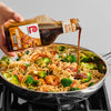 A hand pours Lawrys Honey Teriyaki Marinade over a pan of noodles shrimp and broccoli.